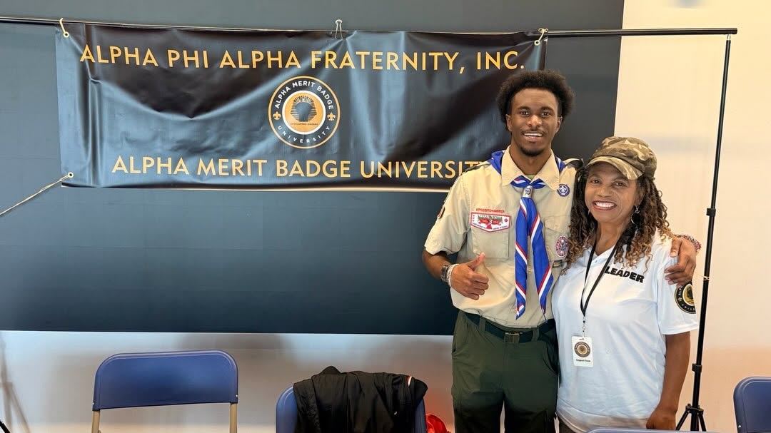 A Scout and a volunteer in front of the Alpha Merit Badge University banner